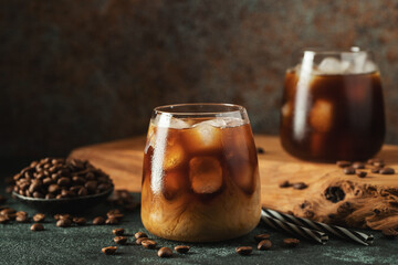 Ice coffee in a tall glass with cream poured over, ice cubes and beans on a dark concrete table.