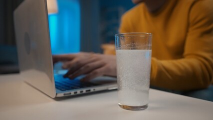 An effervescent tablet dissolves in a glass of water on a table close up. In the background, a man's hands are typing on a laptop. Cure for a cold, pain or hangover. Home medicine concept.