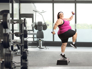 Fototapeta na wymiar Cheerful overweight woman exercising with weights and a stepper at a gym