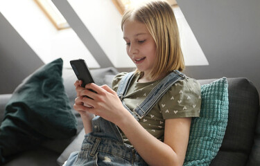Child girl with smartphone sitting on sofa at home and chatting with friend in social media. Pretty preteen kid with mobile phone texting