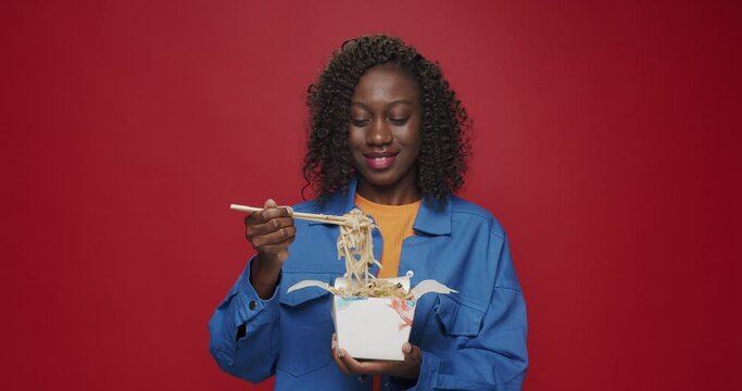 African american woman looking at camera and smiling, preparing to eat noodles, seafood and vegetables with chopsticks on isolated red background. Woman eating Asian noodles for lunch with chopsticks.