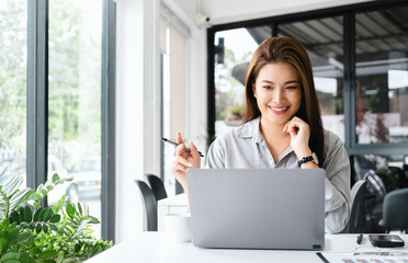 pretty happy young asian entrepreneur woman holding pen working with laptop sitting in the office or cafe.