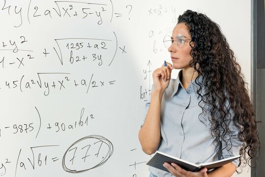 Engaged young Middle Eastern female teacher working on project alone in classroom, looking at whiteboard with math tasks, analyzing notes, thinking over business process schedule, work plan