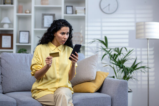 Cheated and sad woman sitting on the sofa inside the house in the living room, Hispanic received a rejection of the transfer, holds a bank credit card in her hands, uses an application on her phone.