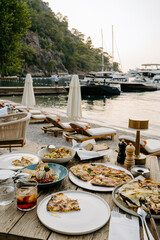 A scenic beachfront breakfast setup with fresh gourmet dishes on a rustic wooden table shaded under lush green foliage. White parasols and lounge chairs overlook the calm blue sea in Türkiye