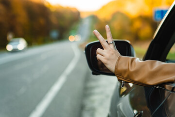 a hand sticks out of the window of a car parked on the side of the road in the autumn season and shows a peace sign