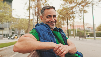 Mature man wearing casual clothes enjoys relaxing on park bench looking at the camera and smiling