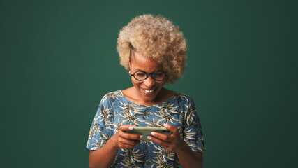 Happy girl in glasses dressed in blue t-shirt, holds mobile phone in her hands, smiles and plays...
