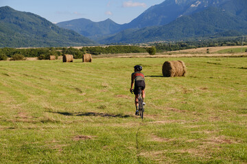 Man cyclist in sports attire, wearing a helmet and glasses, is training on a gravel bike along a gravel road with a mountain view. Muscular guy enjoying his favourite active hobby outdoors.