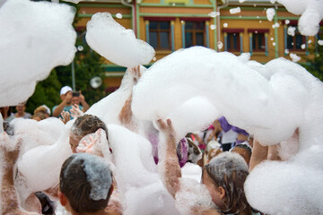 Lot white foam children and adults raise their hands above their heads at summer holiday in water park. Foam party, people have fun raising their hands, catching soap bubbles, summer festival on beach