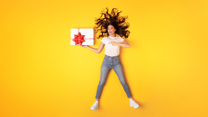 Woman Posing With Gift Pointing At Wrapped Box, Yellow Background