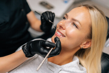 Detail headshot of happy blonde female patient having treatment in dentistry clinic. Close-up hands of unrecognizable dentist in gloves examining patient teeth with dental equipment instrument.