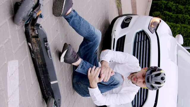Classically dressed man with a protective helmet on his head sitting on the asphalt after falling off his electric scooter following a car accident. Broken knee. Vertical video for story