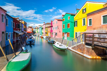 Naklejka na ściany i meble Colourful houses and buildings with reflection in blurred long exposure water of the river. Dock with moored boats on glassy water. Mirror of colors and bright vivid blue sky. Burano, Venice, Italy