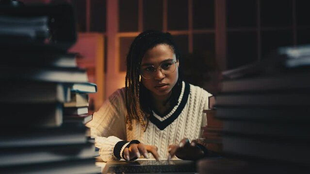 Young black woman typing on laptop surrounded by stacks of books and folders