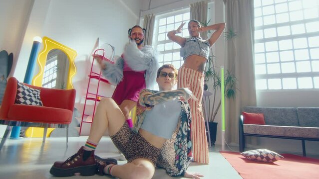 Group of LGBTQ vogue dancers in eye cathing trendy outfits performing dance movements and posing together on camera in studio with trendy designed interior. Low angle, wide shot