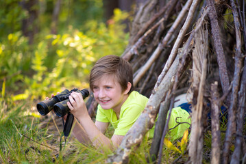 10 year old boy looks through binoculars, lies in a hut made of branches in a summer camp. a happy child spends his summer holidays hiking, outdoors, in the forest.