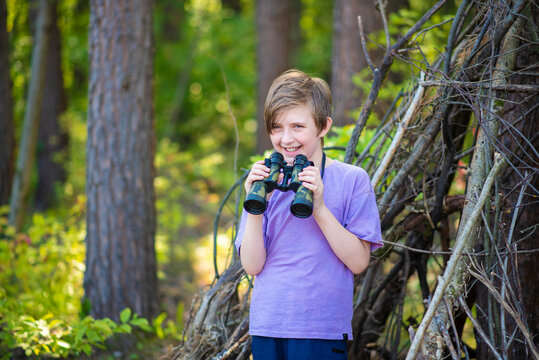 A 10-year-old boy looks through binoculars, stands at a hut made of branches at a summer camp. a happy child spends his summer holidays hiking, outdoors, in the forest. - Powered by Adobe
