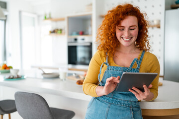 Happy smiling adult woman dressed casually using digital tablet while leaning on table in kitchen....