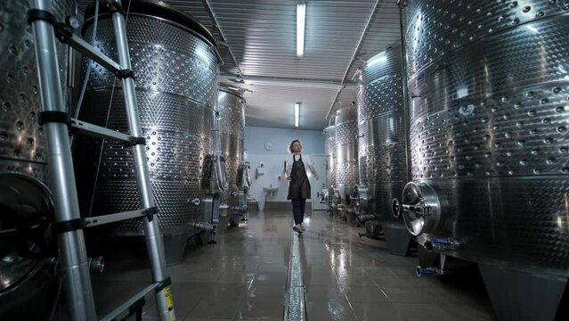 A happy successful farmer or winemaker is walking in the middle of wine tanks and checking a fermentation grade of high quality wine production in a modern winemaking facility or winery.