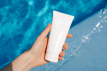 A woman's hand holds a white mockup blank tube of cream on a background of blue pool water. A...