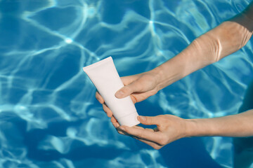 Woman's hands open a white mockup tube of moisturizing cream or sunscreen in blue pool water. Skin...