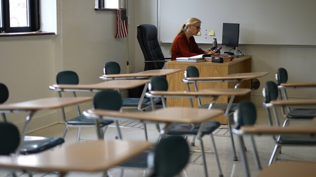 Happy female teacher sitting at desk in empty school classroom desk grading papers working on assignments. US American flag in the background.