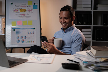 Fototapeta premium Smiling Asian businessman relaxing after work hard in the office, holding smartphone and coffee cup at office desk.