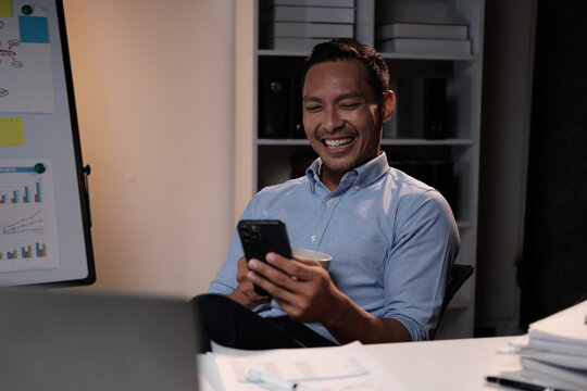 Smiling Asian businessman relaxing after work hard in the office, holding smartphone and coffee cup at office desk.