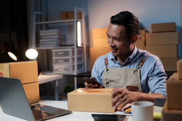 Fototapeta premium Young smiling hardworking small business owner working in his workplace while using smartphone, working during the night.