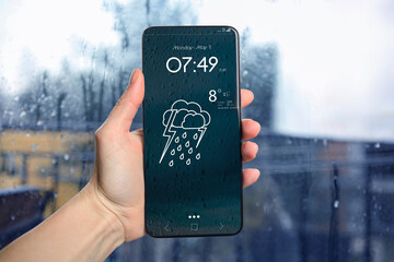 Woman checking weather using app on smartphone near wet window, closeup. Data and illustration of...