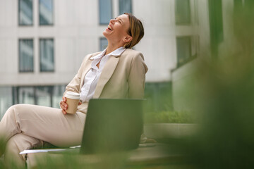 Smiling business woman sitting on bench and drinking coffee during break time near office building