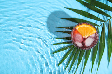 Glass of cold Negroni cocktail and palm leaf in water on color background