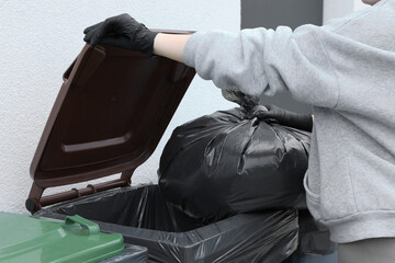 Woman throwing trash bag full of garbage in bin outdoors, closeup. Recycling concept
