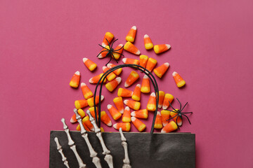 Shopping bag with tasty candy corns, Halloween decor and skeleton hand on pink background
