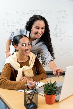 Tutor explaining research specific to student in library. Two Oriental woman in casual sitting and standing at desk, using laptop, pointing at monitor and talking. Consulting concept