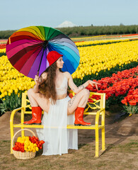 Woman in white dress and red boots with rainbow umbrella on yellow bench before field of red and yellow tulips