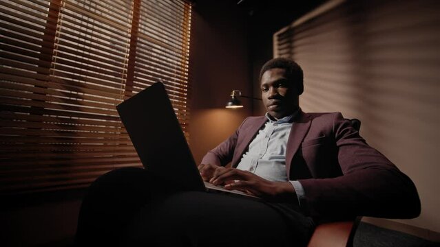 African American worker typing on laptop message or text to his work partner while sitting in armchair in shadow room Actively working in quiet place with closed blinds and lamp with bright light.