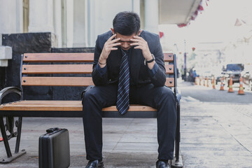 Sad failed young Asian businessman sitting alone on bench touching his head feeling despair