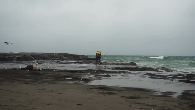 A man is fishing on the muriwai beach which is quite windy