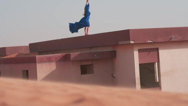 A young woman with her hair flowing in the wind and wearing a long blue cloak stands barefoot on the roof of a sand-covered house in the middle of the desert