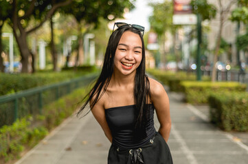 A pretty young lady standing in a park wearing a black strapless tank top and sunglasses, leans forward smiling widely while hands are behind her back. Trees,bushes and fence are in the background.