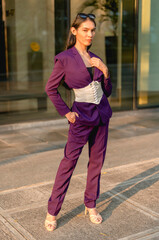 An asian lady standing outside the office wearing a purple suit and purple slacks poses with a cool and confident expression. A window glass in the background.
