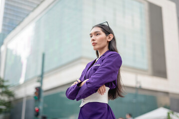 A lovely lady standing outside in a purple suit and purple slacks does an arms crossed pose, looking serious while glancing far in front. Buildings, tents and people in the background.