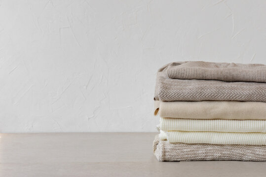 Wool knitted clothes stack closeup in neutral beige colors on table on white empty wall background. Aesthetic fashion minimalist wardrobe concept, warm autumn knitwear jersey