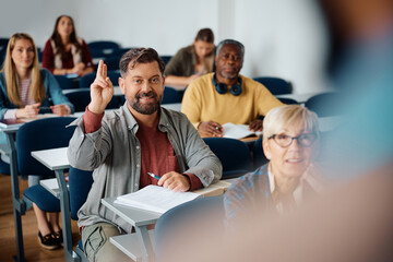 Mature man raising hand to ask question while attending class in lecture hall. - obrazy, fototapety, plakaty