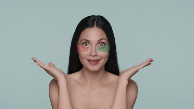 Portrait of a seminude woman with various hyaluronic patches under her eyes on a blue background close up. The woman taps the patches with her fingers, smoothing them out. Slow motion.