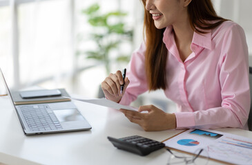 Asian businesswoman using calculator to calculate turnover Company earnings on graph paper data, charts, reports working on laptop computer lying on table for meeting preparation discuss in the office