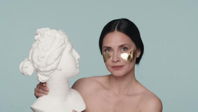 Profile portrait of a seminude woman with golden patches under her eyes, holding a plaster bust in her hands. The woman turns her face and nods her head in the affirmative. Slow motion.