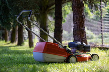 Lawn mower set in the shade of coniferous trees. Delicately blurred background, shot on a sunny day.
